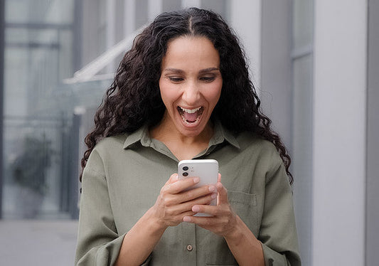 Excited woman with curly hair looking at her smartphone outdoors, smiling and celebrating a happy moment.