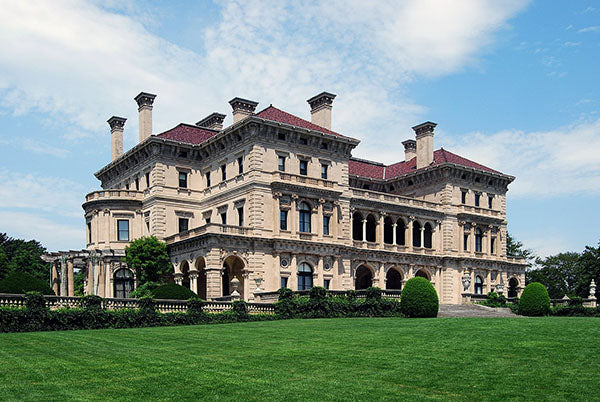 A grand mansion with classical architecture, featuring large columns, multiple balconies, and a well-maintained lawn, under a clear blue sky.
