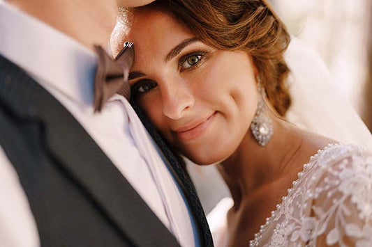 Close-up portrait of a bride smiling with dimples while leaning against her groom. She is wearing elegant bridal earrings and a lace wedding dress, capturing a tender and intimate moment.