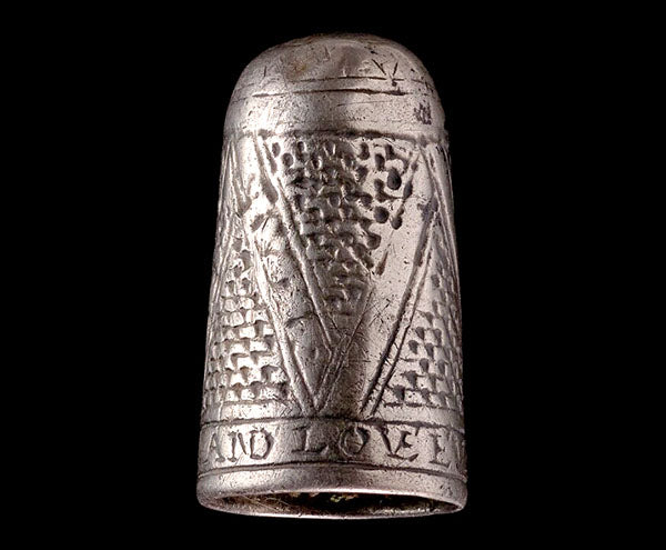 An antique silver thimble featuring intricate engraved patterns, including a symbolic letter 'V' and the inscription 'AND LOVE,' displayed against a black background.