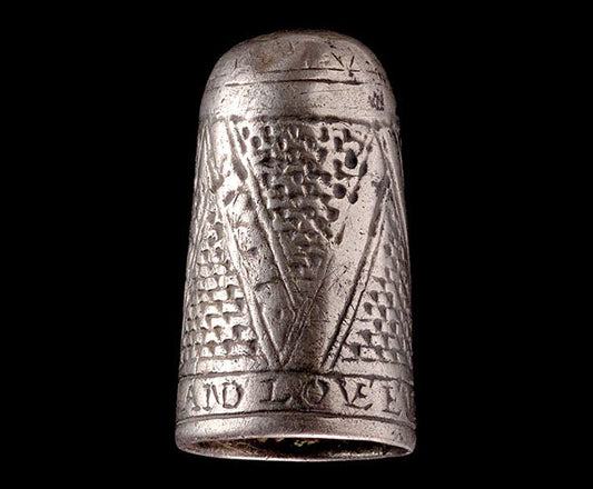 An antique silver thimble featuring intricate engraved patterns, including a symbolic letter 'V' and the inscription 'AND LOVE,' displayed against a black background.