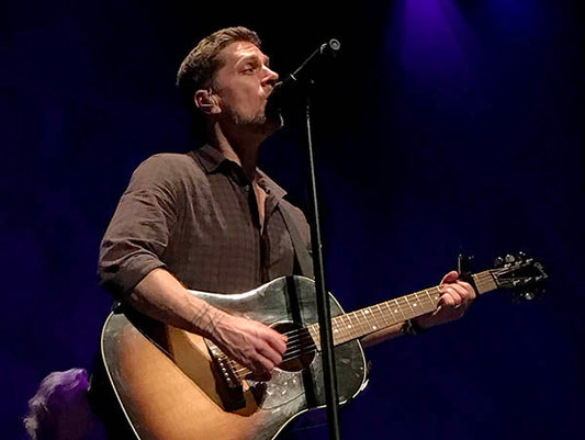 Musician performing on stage with a guitar, singing into a microphone under purple stage lights.