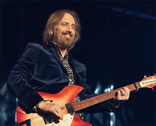 Legendary musician Tom Petty performing on stage with a guitar, smiling and enjoying the moment. He is wearing a blue velvet jacket and a patterned shirt, with a spotlight highlighting his presence during the live concert.