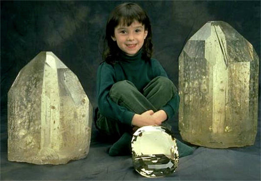 Young girl sitting with large, clear crystals on either side of her, holding a smaller crystal in front, with a backdrop of dark fabric.