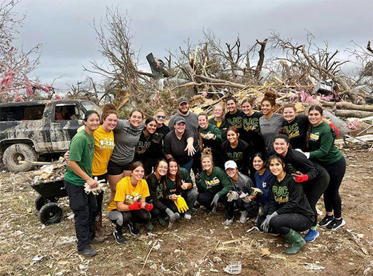 Group of volunteers standing together in front of debris from a tornado, with some showing support and smiling, while one person is holding up an engagement ring.