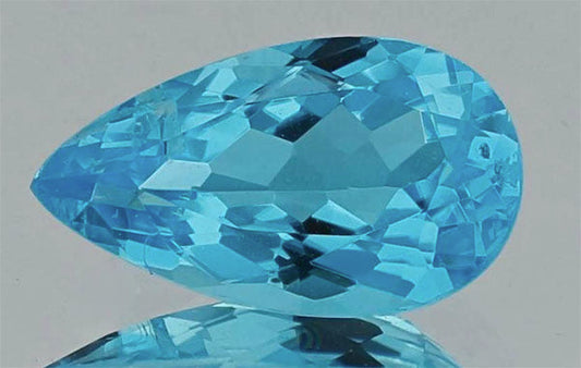 Close-up of a stunning blue topaz gemstone with a pear shape, showcasing its brilliant facets and vivid blue color. The gemstone is placed on a reflective surface, highlighting its clarity and shine.