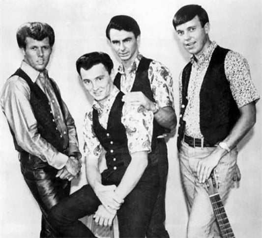 Black and white photo of a 1960s rock band with four members dressed in floral shirts and vests, posing together with one holding a guitar.