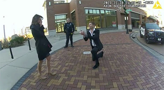 Man proposing to his girlfriend in the street while she looks surprised, with two police officers and a squad car visible in the background.