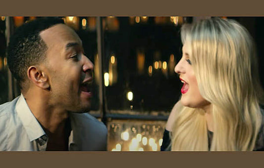 John Legend and Meghan Trainor singing together in a duet with blurred lights in the background.
