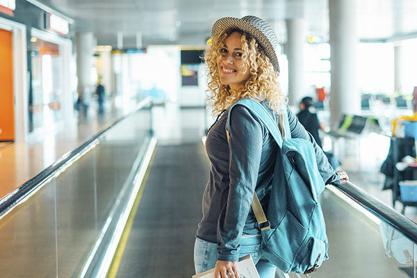 Smiling young woman with curly hair wearing a hat and backpack, walking through an airport terminal with a relaxed and joyful expression.