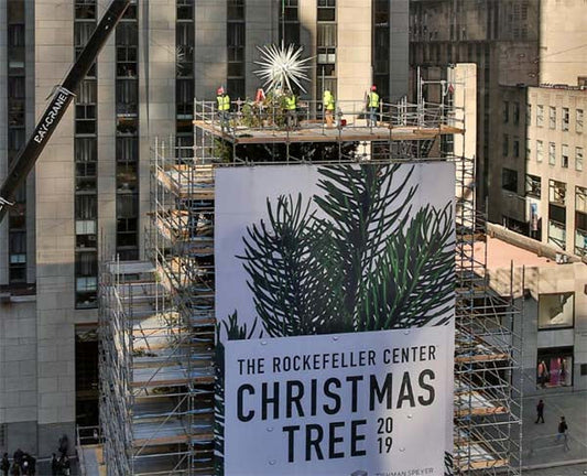 Workers placing the star on top of the Rockefeller Center Christmas tree in 2019, with a large billboard showcasing the tree.