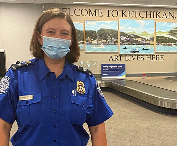 Female TSA officer in uniform wearing a mask stands in front of a mural at Ketchikan airport with the words 'Welcome to Ketchikan, Art Lives Here' in the background.