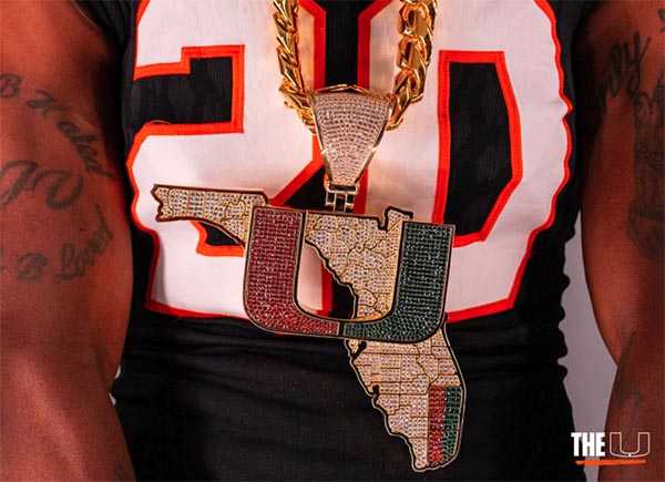 Close-up of a person wearing a large, flashy pendant shaped like the state of Florida with the 'U' logo, encrusted with diamonds and colorful gems. The pendant is part of a gold chain, and the person is dressed in a black sports jersey with the number 20.