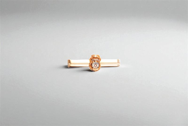 Gold tie bar with a badge-style emblem centered on a light gray background, minimal product photo.