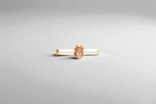 Gold tie bar with a badge-style emblem centered on a light gray background, minimal product photo.
