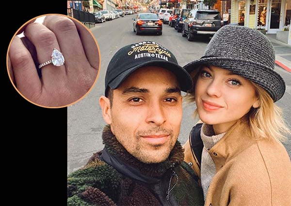 Couple posing for a selfie on a street, with the woman showing her engagement ring featuring a pear-shaped diamond, and the man wearing a cap.