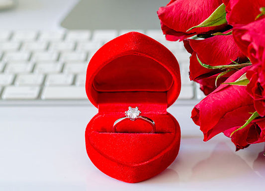 Diamond engagement ring in a red velvet box with fresh red roses in the background, set on a white keyboard.