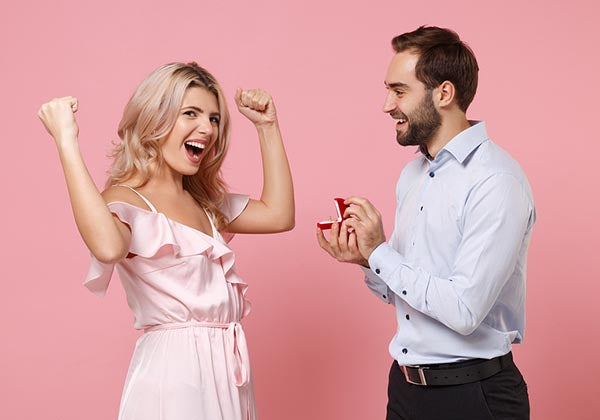 Happy woman celebrating with her arms raised as her partner proposes with a ring in a red box against a pink background.