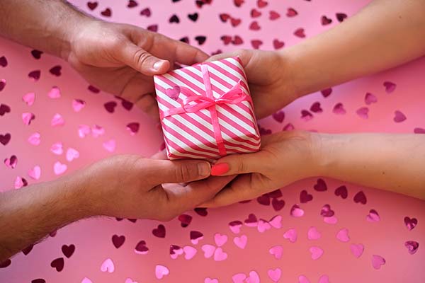 Close-up of two hands exchanging a pink and white striped gift box with a pink ribbon, set against a pink background covered in heart-shaped confetti. The image conveys a romantic and celebratory vibe.