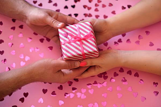 Close-up of two hands exchanging a pink and white striped gift box with a pink ribbon, set against a pink background covered in heart-shaped confetti. The image conveys a romantic and celebratory vibe.