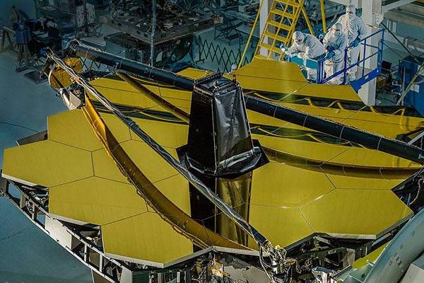 Close-up of the James Webb Space Telescope during assembly, showing the golden hexagonal mirror segments. Engineers in protective suits work on the telescope, with a ladder and equipment in the background.