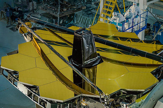 Close-up of the James Webb Space Telescope during assembly, showing the golden hexagonal mirror segments. Engineers in protective suits work on the telescope, with a ladder and equipment in the background.