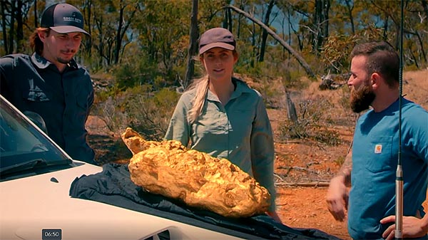 Three people standing beside a car, admiring a large gold nugget placed on a cloth in the Australian outback.