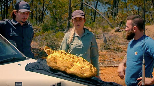 Three people standing beside a car, admiring a large gold nugget placed on a cloth in the Australian outback.