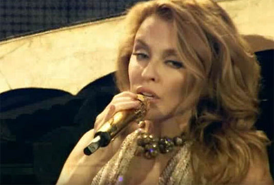 Kylie Minogue performing live on stage, holding a golden microphone and wearing a glamorous outfit with a statement necklace.