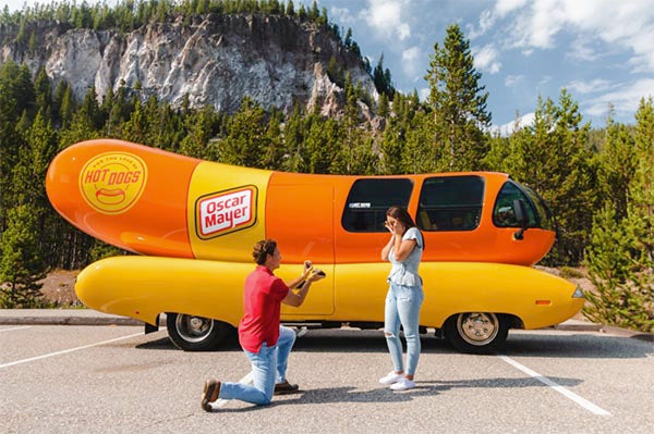 Man proposing to a woman in front of the iconic Oscar Mayer Wienermobile, a large hot dog-shaped vehicle. The woman is surprised and emotional, while the man is kneeling with a ring, in a scenic outdoor setting with mountains in the background.