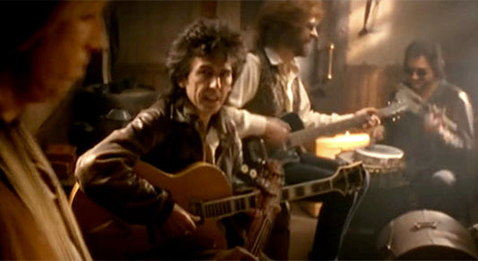 Group of musicians playing instruments in a dimly lit room. One man plays a guitar while another plays a banjo, and two others are playing percussion instruments, creating a cozy, intimate jam session.