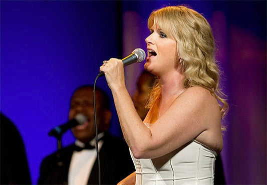 Female singer performing on stage with a microphone, wearing a white dress, with background singers visible.