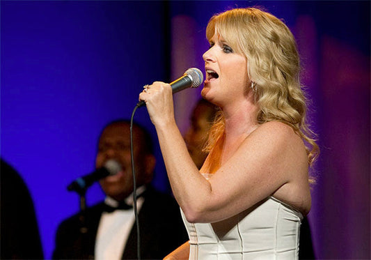 Country music singer performing on stage, holding a microphone with passion, wearing a white strapless dress, with a backup vocalist visible in the background.