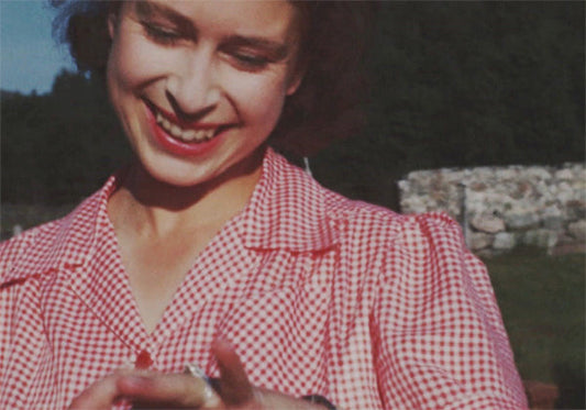 Smiling woman in red checkered shirt looking at ring on her finger outdoors with stone wall in background.