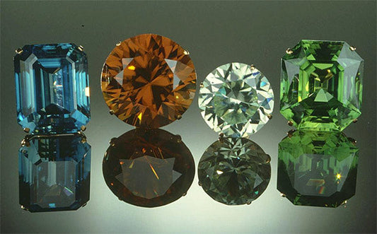 Collection of colorful gemstones, including blue, orange, clear, and green stones, displayed in different cuts such as emerald and round, with reflections on the surface.