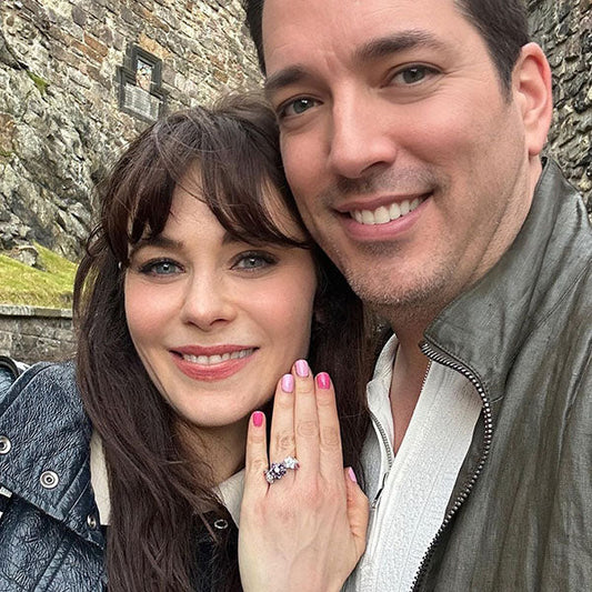 Zooey Deschanel and Jonathan Scott posing for a selfie, showing off Zooey's engagement ring with a purple gemstone in front of a scenic background.